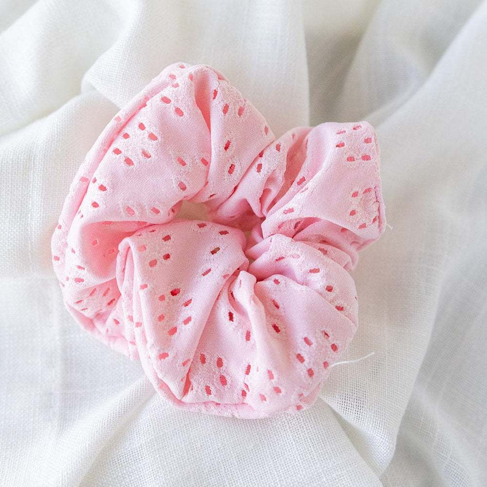 A petal pink eyelet lace scrunchie with delicate details rests on white cloth, offering a charming hair accessory for any occasion.