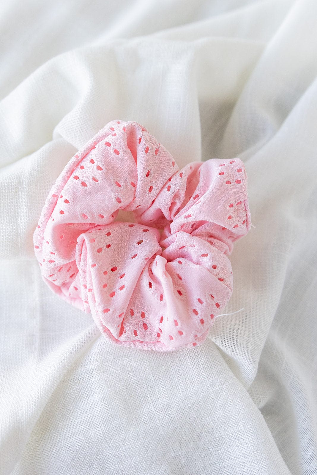 A petal pink eyelet lace scrunchie with delicate details rests on white cloth, offering a charming hair accessory for any occasion.