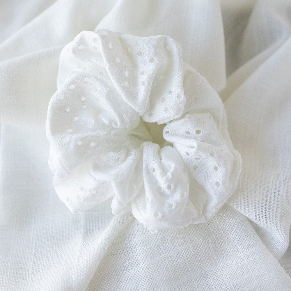 A white Eyelet Lace Scrunchie with delicate patterns is elegantly placed on a matching textured cloth background, perfect as a versatile hairstyle accessory.