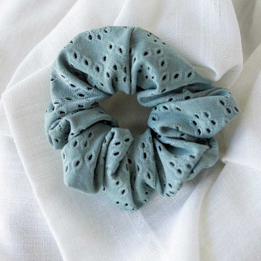 A chic ponytail is perfectly complemented by a sage green eyelet lace scrunchie resting on white fabric.