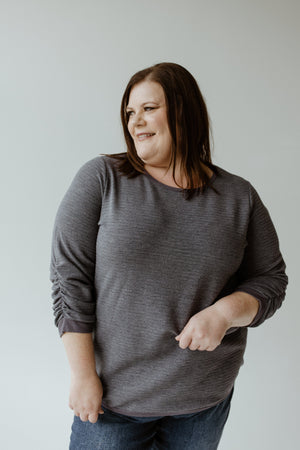 A person wearing a gray sweater and jeans stands against a plain background, looking to the side with a smile, as if considering the allure of Yesta's 3/4 Ruched Sleeve Shimmer Top in Night Shadow.