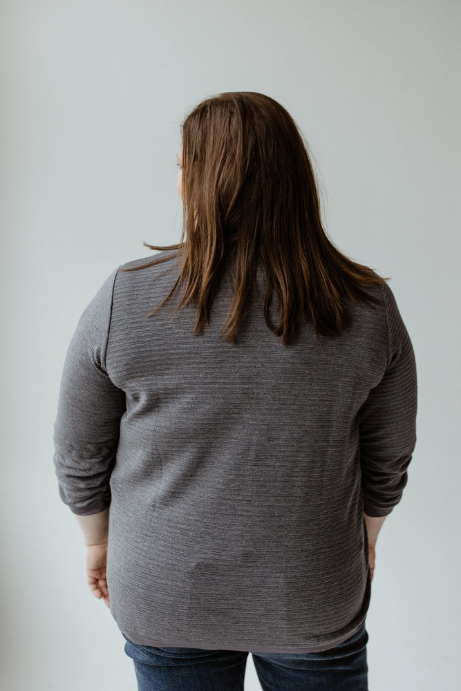 A person with long brown hair, seen from behind against a plain backdrop, is wearing jeans and the Yesta 3/4 Ruched Sleeve Shimmer Top in Night Shadow.