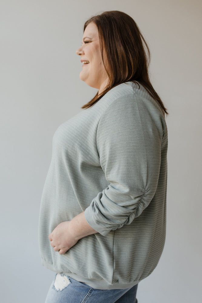 A person with shoulder-length brown hair wearing the Yesta 3/4 Ruched Sleeve Shimmer Top in Quietude and jeans stands with their back to the camera against a neutral background.