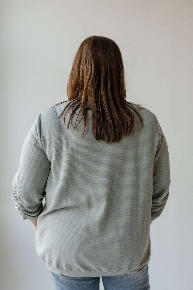 A person with shoulder-length brown hair wearing the Yesta 3/4 Ruched Sleeve Shimmer Top in Quietude and jeans stands with their back to the camera against a neutral background.