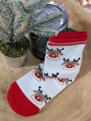 A pair of festive reindeer socks with a Rudolph design sits next to a small potted evergreen tree on a cozy white background, adding charm to the wooden surface.