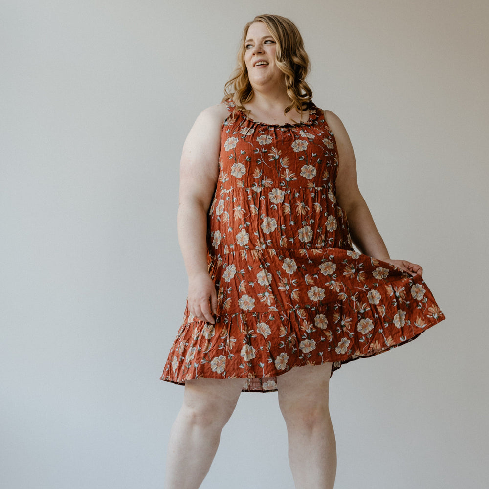 Wearing the RUFFLE NECKLINE TIERED KNEE LENGTH DRESS IN EMBER by Mikarose, a person smiles and strikes a pose in a studio setting.