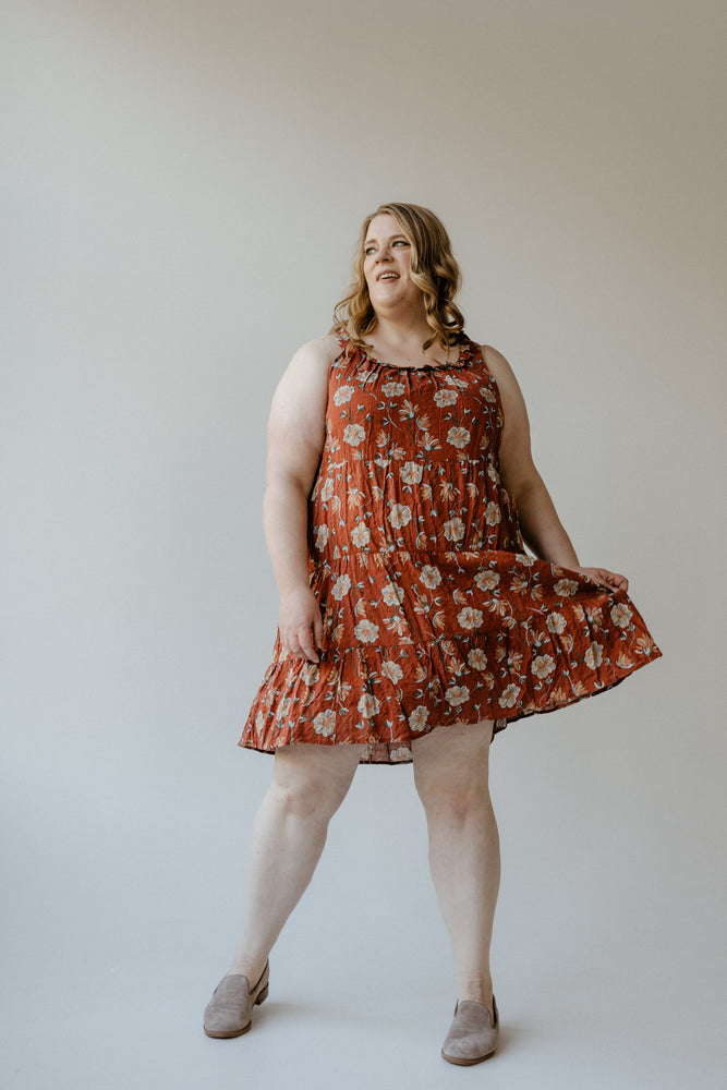 Wearing the RUFFLE NECKLINE TIERED KNEE LENGTH DRESS IN EMBER by Mikarose, a person smiles and strikes a pose in a studio setting.