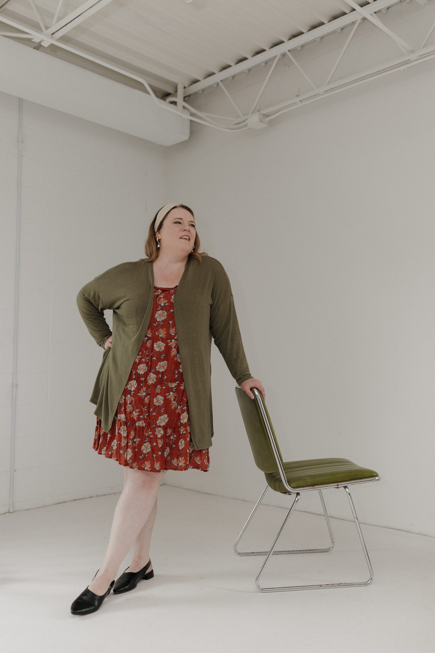 Woman in a red floral dress and green cardigan standing next to a green chair in a white room.