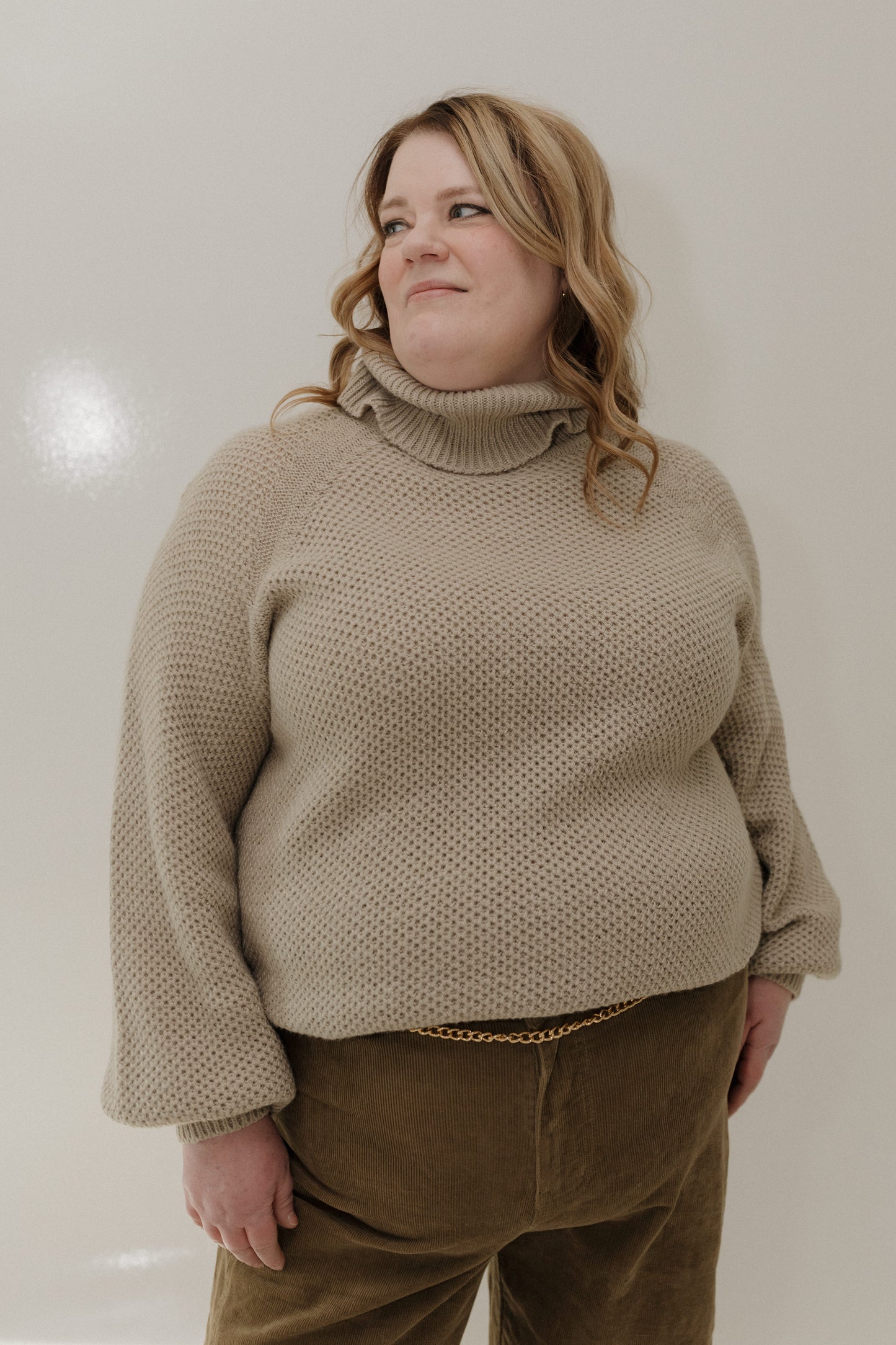 Person wearing a beige knitted sweater and brown pants against a plain background