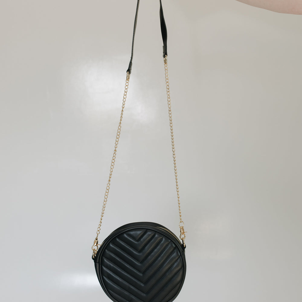 Woman holding the round quilted cross-body purse in black while she is wearing a beige top and black skirt, standing against a tiled wall.