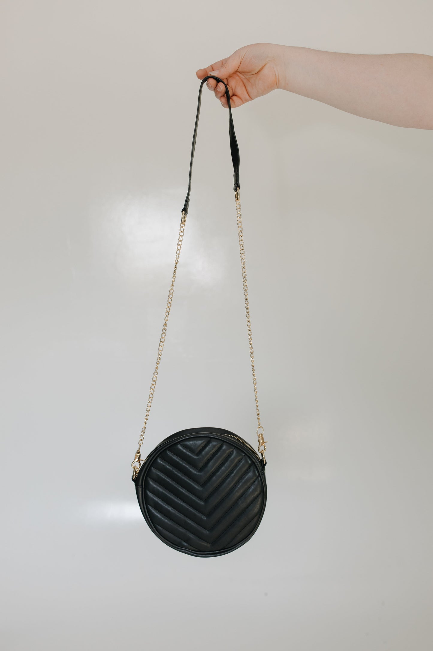 Woman holding the round quilted cross-body purse in black while she is wearing a beige top and black skirt, standing against a tiled wall.