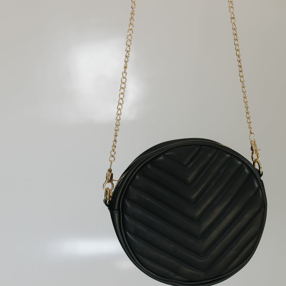 Woman holding the round quilted cross-body purse in black while she is wearing a beige top and black skirt, standing against a tiled wall.