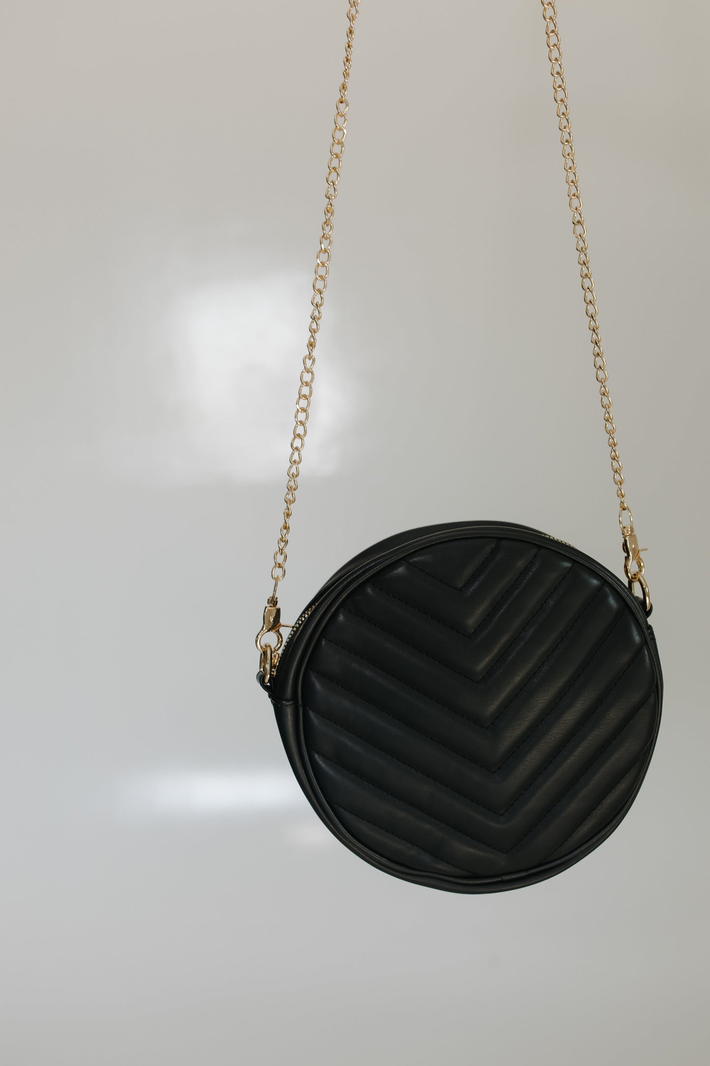 Woman holding the round quilted cross-body purse in black while she is wearing a beige top and black skirt, standing against a tiled wall.