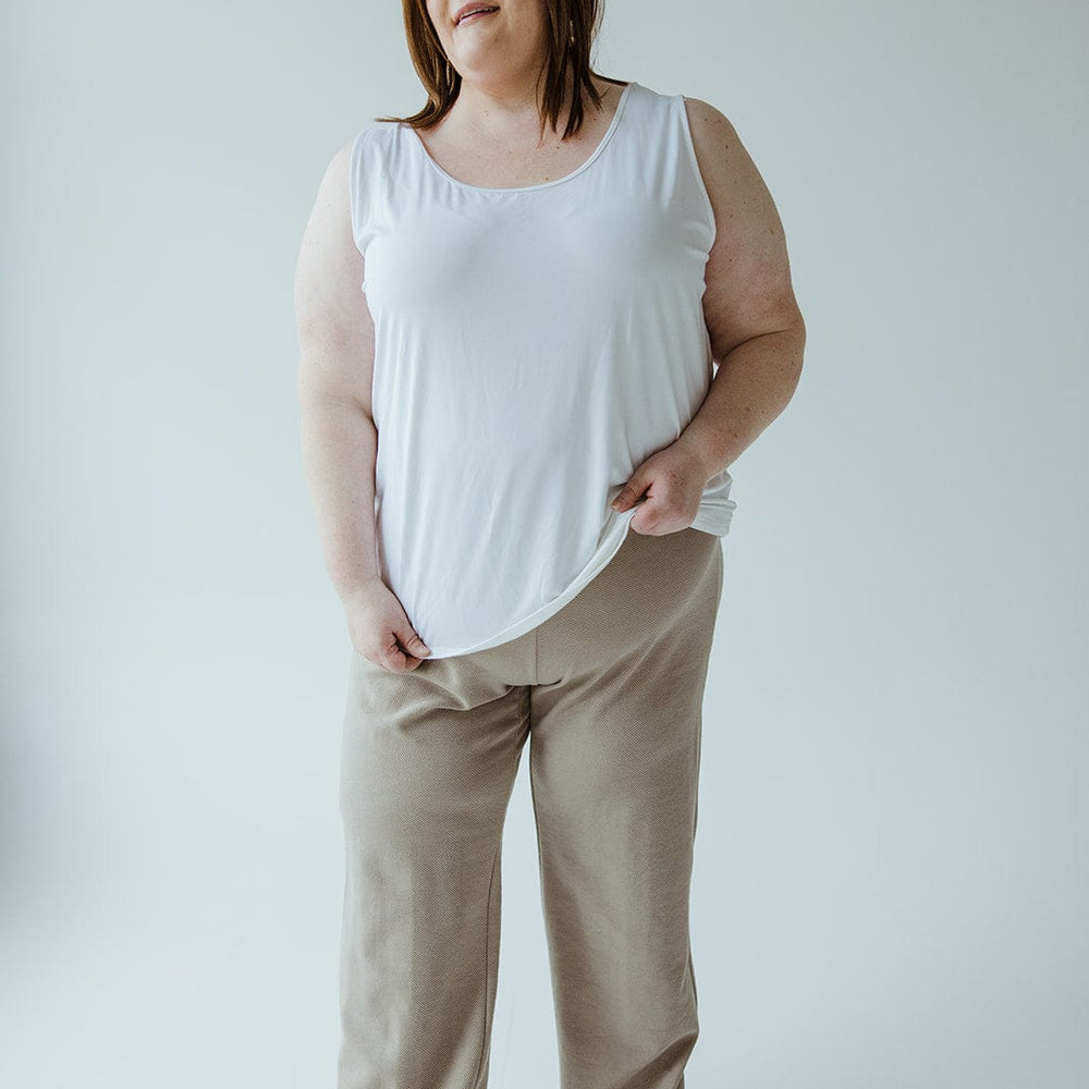 A person stands against a plain background, wearing a white sleeveless top, Yesta's Light Khaki Straight Leg Pull-On Pant made from lightweight textured fabric, and light-colored shoes. They have their hands at their sides and are looking slightly off-camera.