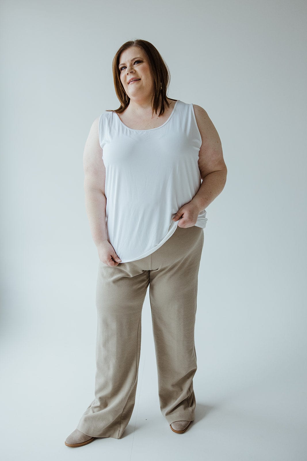 A person stands against a plain background, wearing a white sleeveless top, Yesta's Light Khaki Straight Leg Pull-On Pant made from lightweight textured fabric, and light-colored shoes. They have their hands at their sides and are looking slightly off-camera.