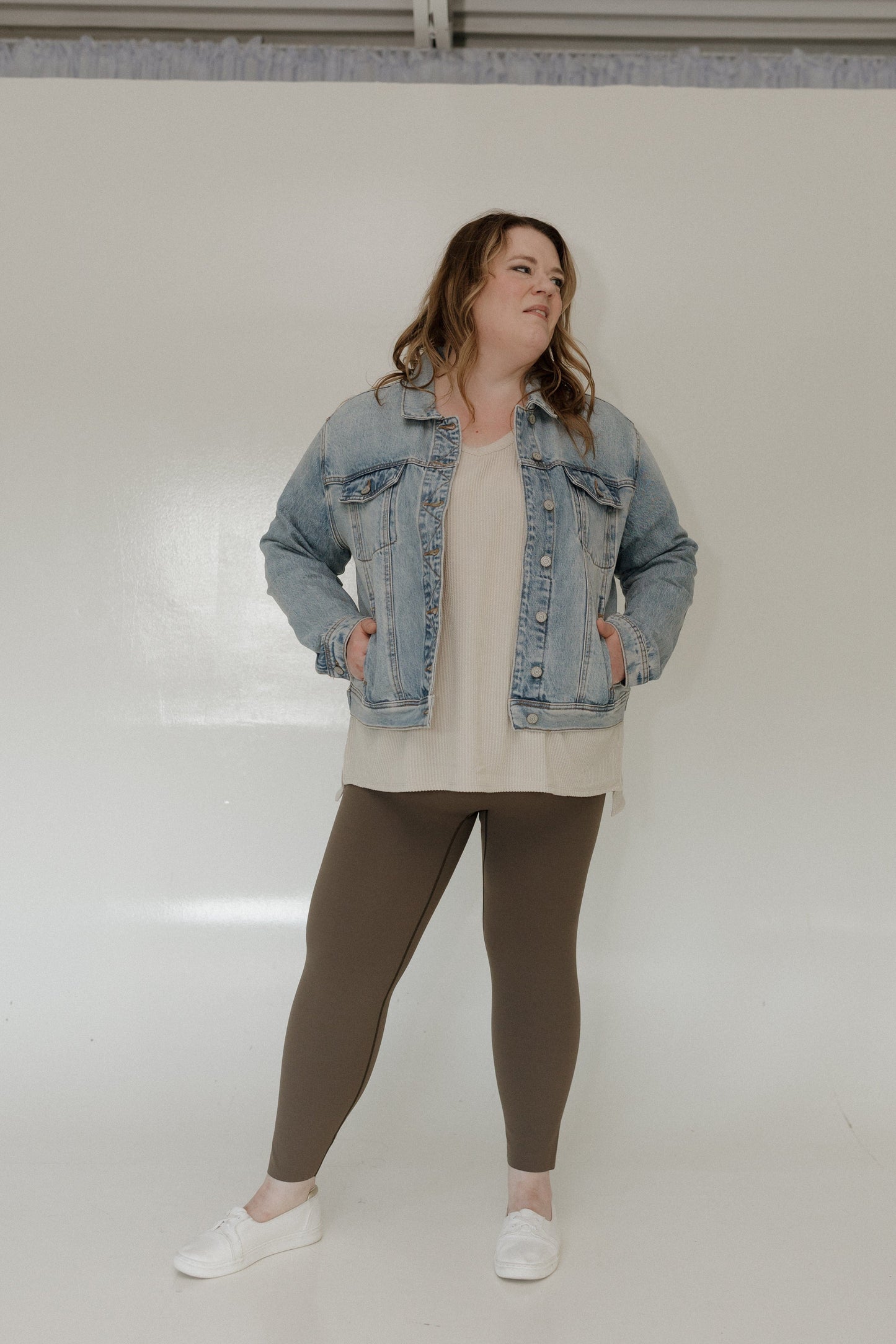 Woman wearing a denim jacket, sand top, and brown leggings against a white background