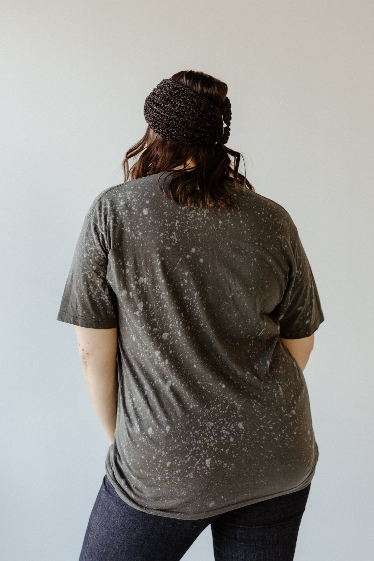 Rear view of a person with long hair wearing a black headband, a hand-bleached funny graphic tee, and dark jeans, standing against a plain gray background.