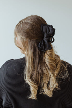 A woman with wavy, blonde hair styled in Darling Effect's Satin Bow Hair Claw in Black looks away from the camera, wearing a dark top.