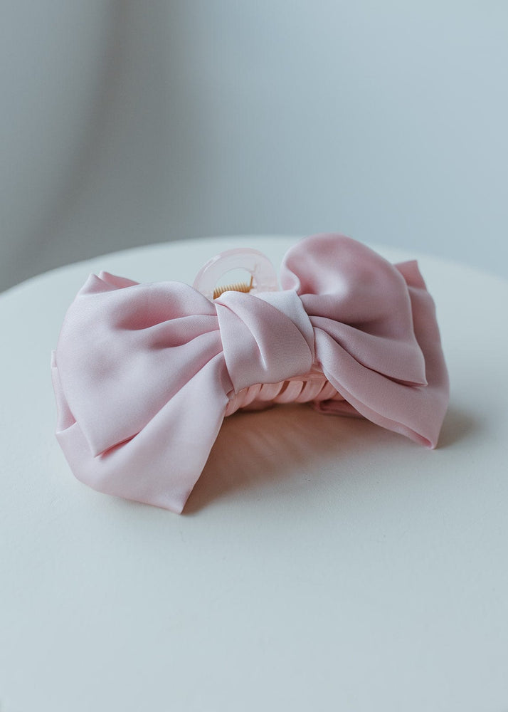 The SATIN BOW HAIR CLAW IN BALLERINA by Love Marlow lies elegantly on a white, curved table surface, making for the perfect hair accessory.