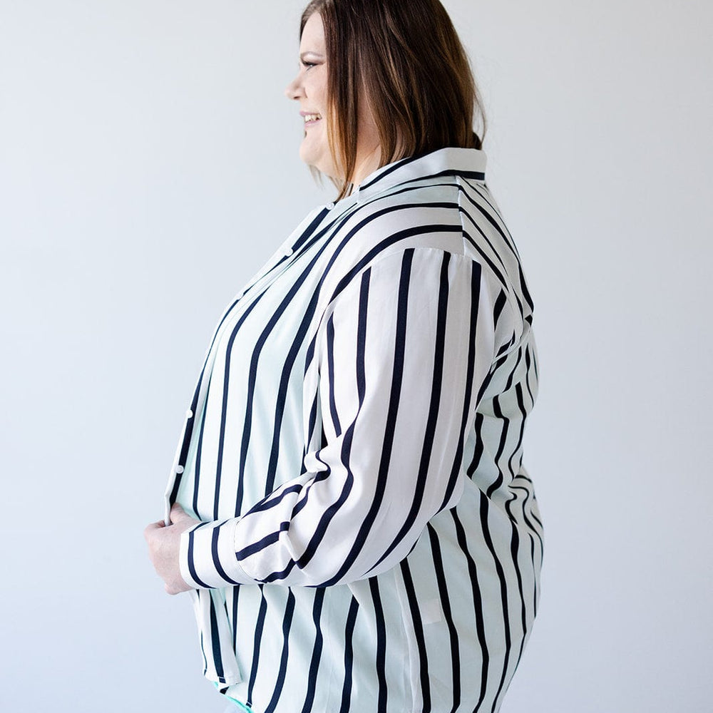 A person stands in profile against a plain backdrop, exuding chic sophistication while wearing Love Marlow's luxurious Satiny Button-Up Blouse in Silky White and Navy Stripe, paired with light blue jeans.