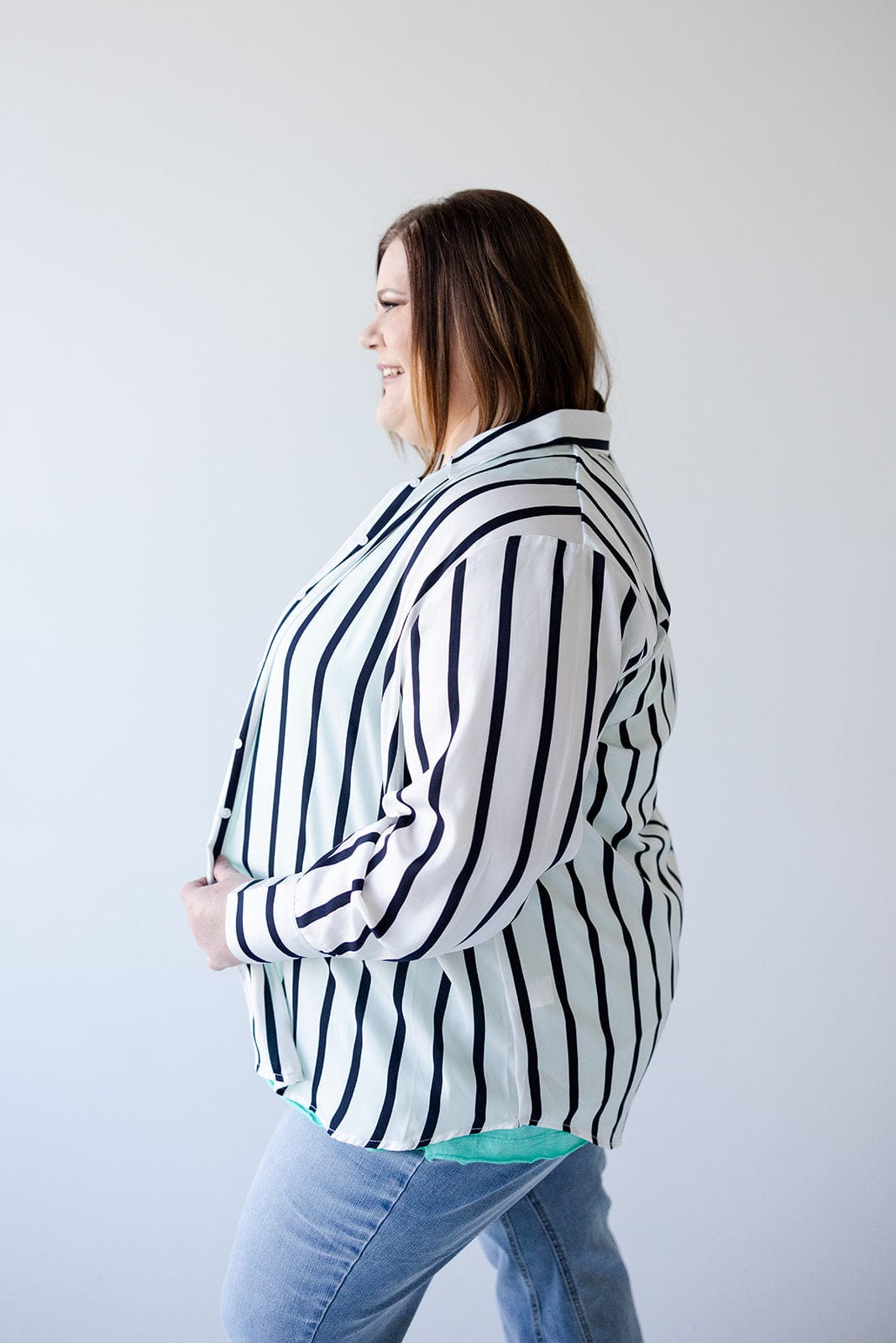 A person stands in profile against a plain backdrop, exuding chic sophistication while wearing Love Marlow's luxurious Satiny Button-Up Blouse in Silky White and Navy Stripe, paired with light blue jeans.