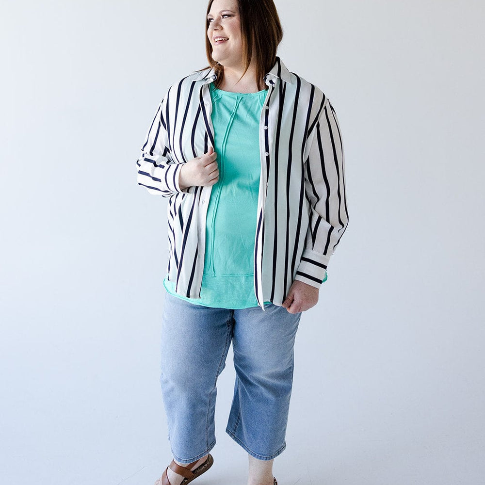 A person stands against a plain background, exuding chic sophistication in the Love Marlow SATINY BUTTON-UP BLOUSE IN SILKY WHITE AND NAVY STRIPE. The ensemble is completed with a turquoise shirt underneath, cropped jeans, and sandals.