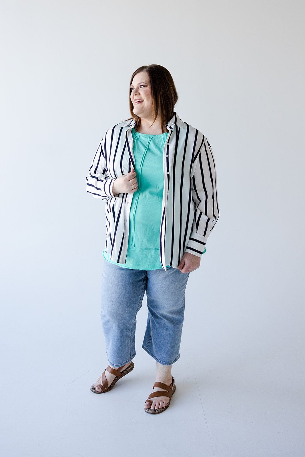 A person stands against a plain background, exuding chic sophistication in the Love Marlow SATINY BUTTON-UP BLOUSE IN SILKY WHITE AND NAVY STRIPE. The ensemble is completed with a turquoise shirt underneath, cropped jeans, and sandals.