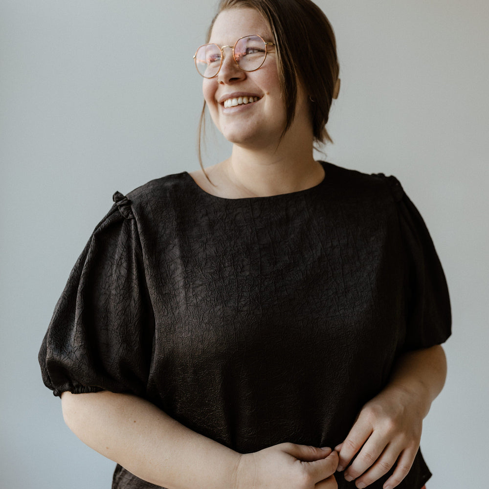 A person wearing glasses, a black blouse, and red pants stands smiling against a plain background.