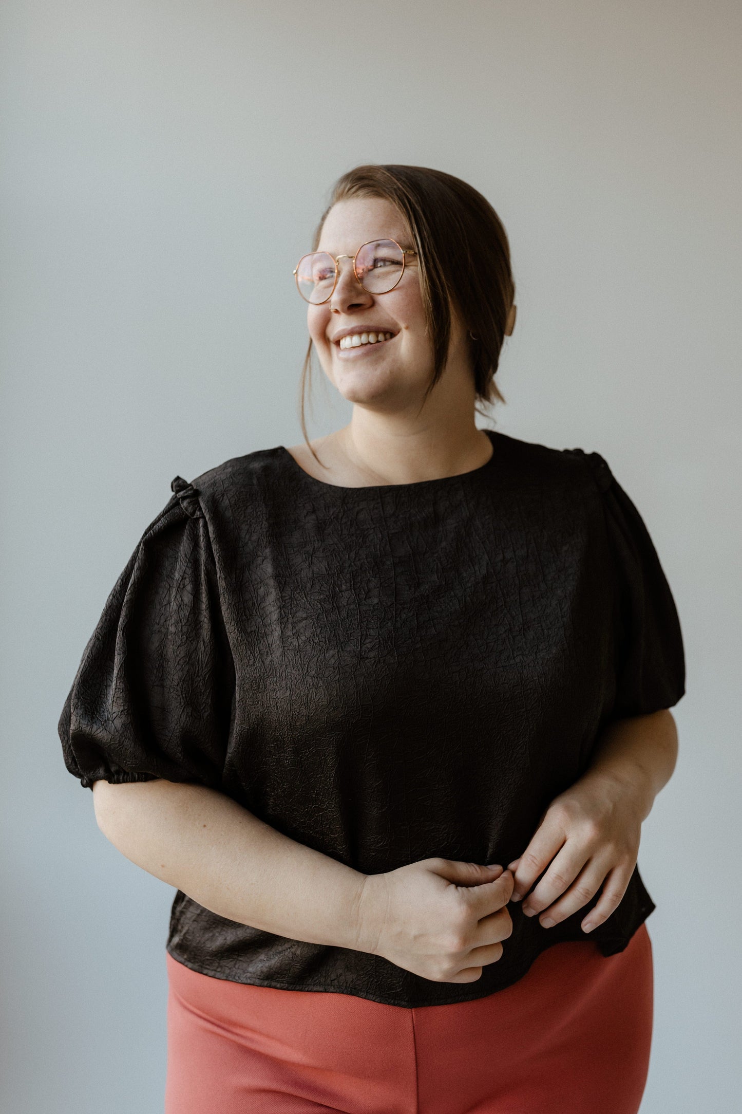 A person wearing glasses, a black blouse, and red pants stands smiling against a plain background.