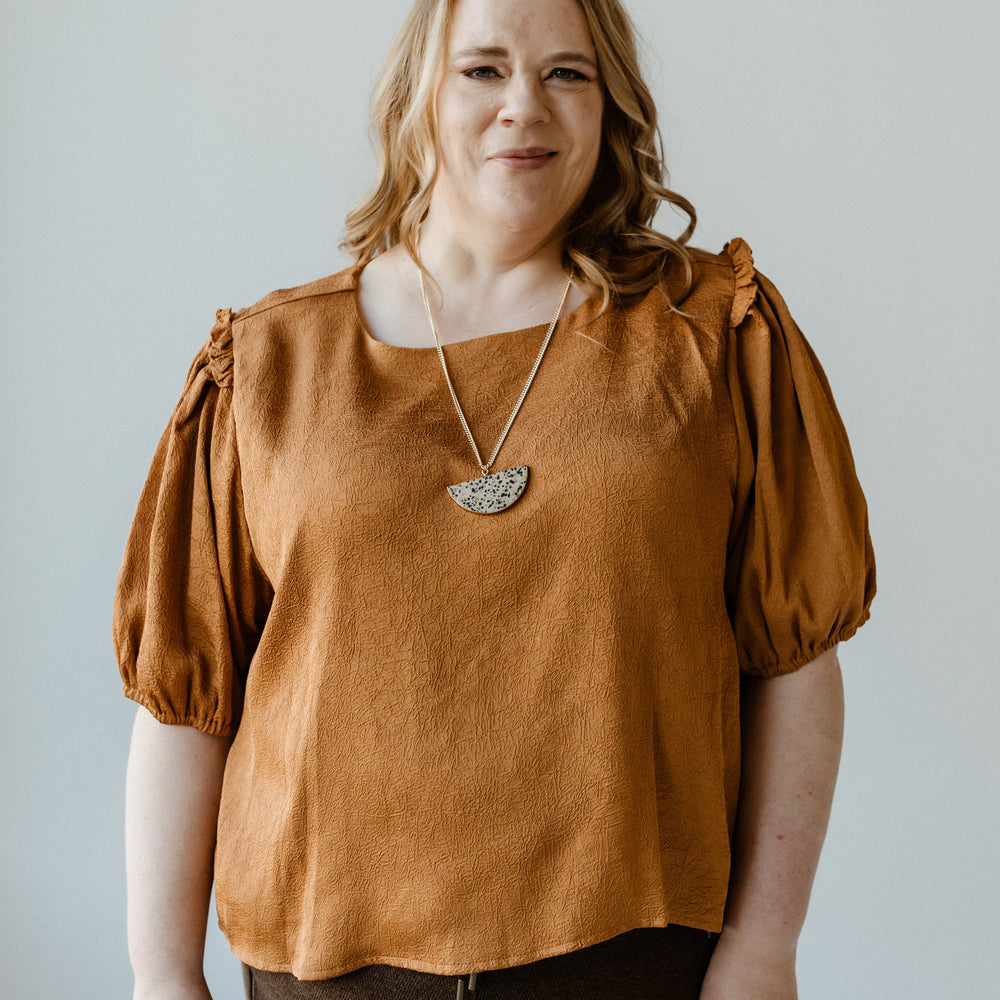 A person with long hair wearing a butterscotch satiny crinkle blouse with ruffled puff sleeves and dark pants stands in front of a light-colored background.