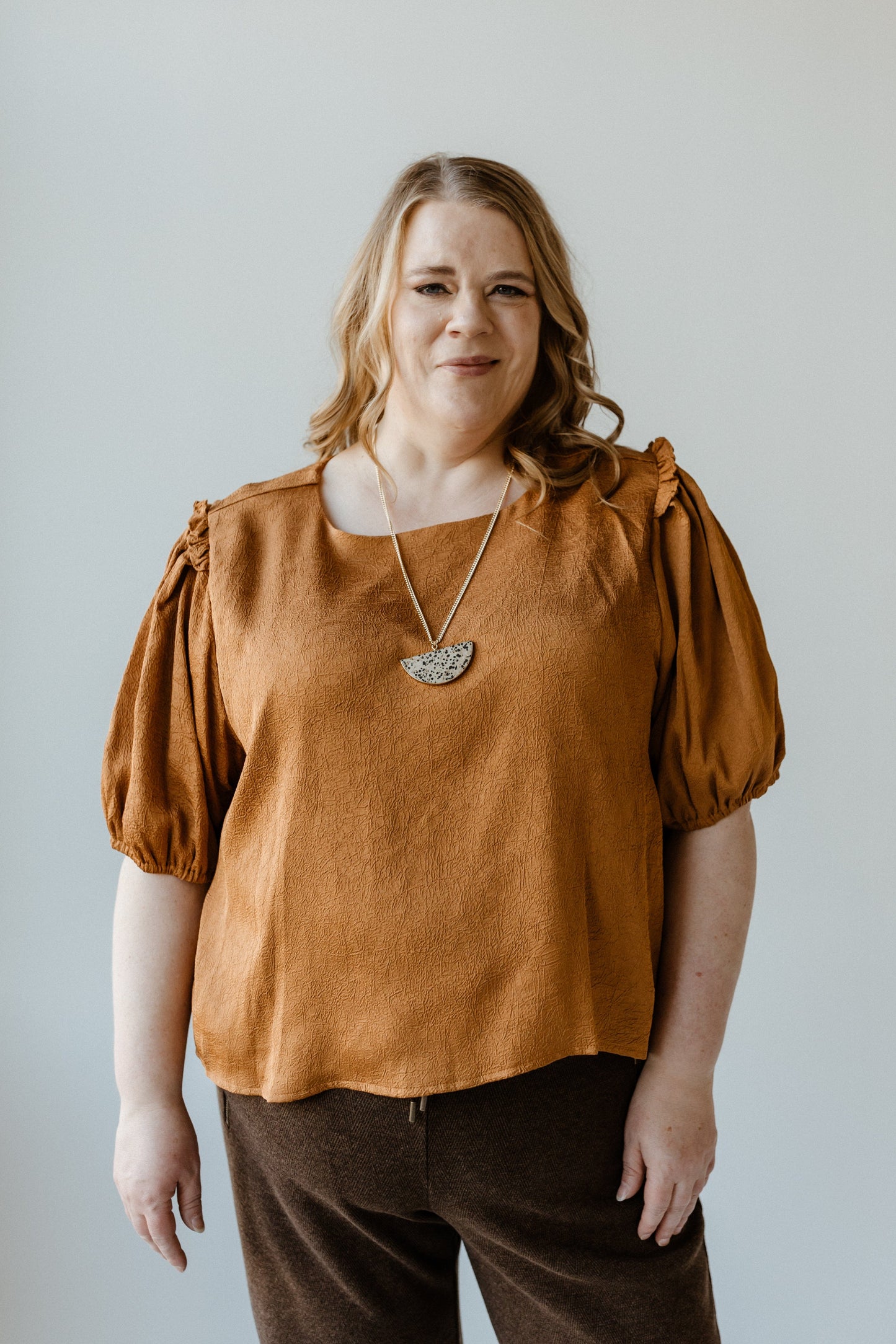 A person with long hair wearing a butterscotch satiny crinkle blouse with ruffled puff sleeves and dark pants stands in front of a light-colored background.