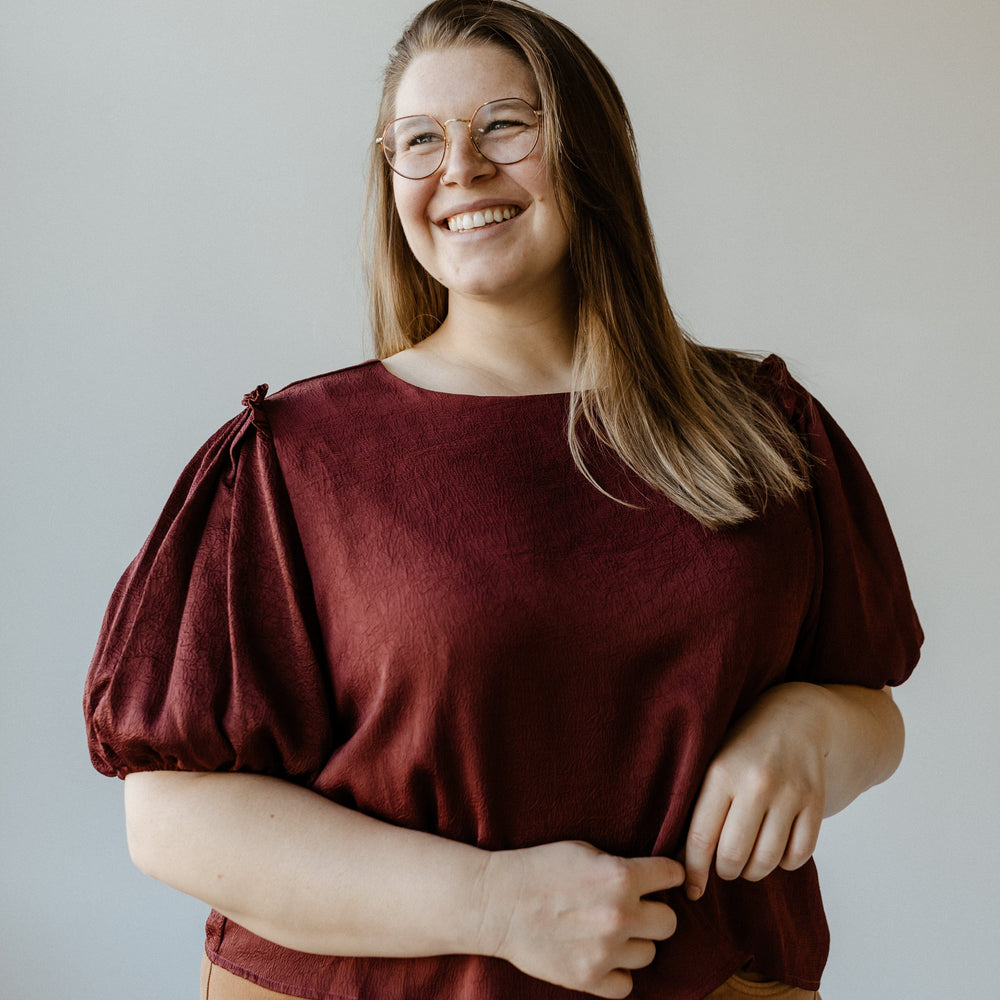 A person with long hair and glasses smiles while wearing a Merlot satiny crinkle blouse and brown pants against a plain background.