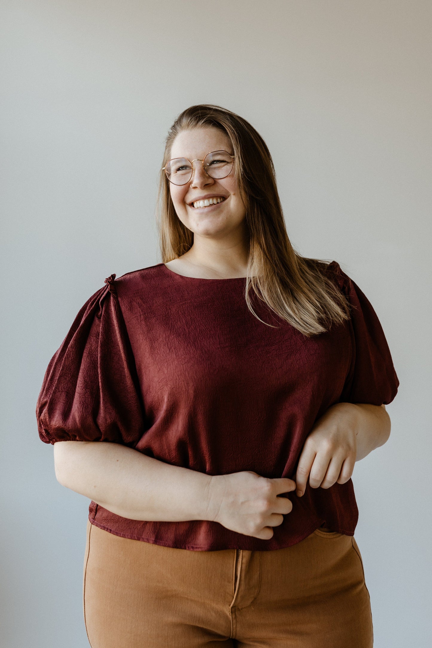 A person with long hair and glasses smiles while wearing a Merlot satiny crinkle blouse and brown pants against a plain background.