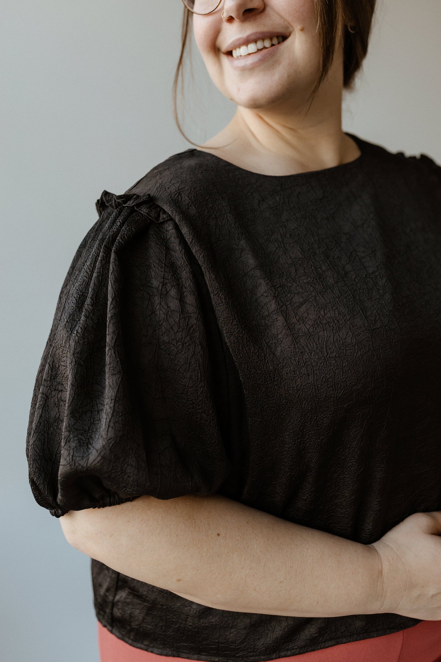 A person in a black blouse with ruffled puff sleeves and a red skirt smiles slightly while looking off-camera.