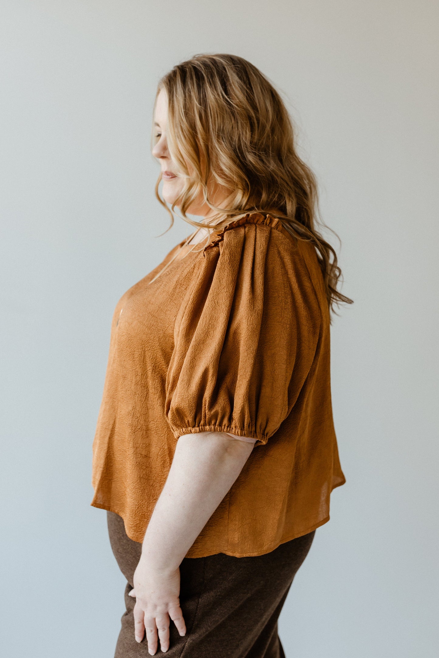 A person with long, wavy hair wearing a brown Butterscotch Satiny Crinkle Blouse, featuring ruffled puff sleeves, is facing slightly to the side against a plain background.