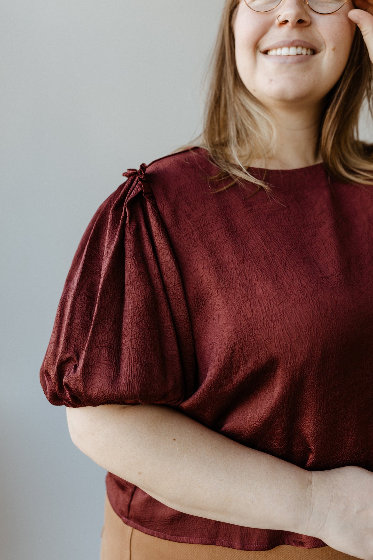 A person in a Merlot satiny crinkle blouse touches their glasses while smiling.