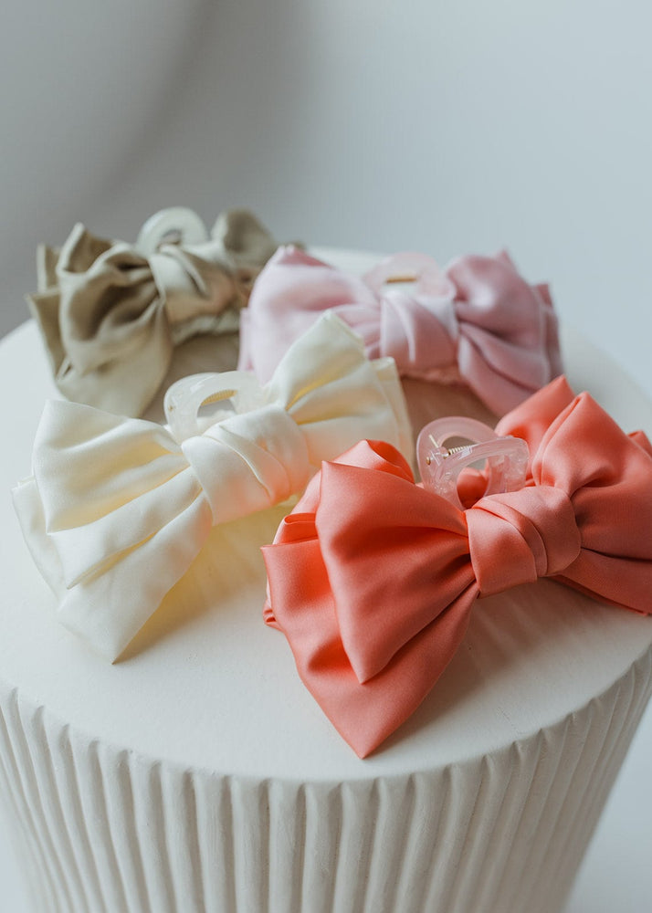 A collection of hair bows and scrunchies in blush pink, beige, white, and coral, featuring the stunning Love Marlow Satin Bow Hair Claw in Blush Pink, all beautifully displayed on a round white textured surface.