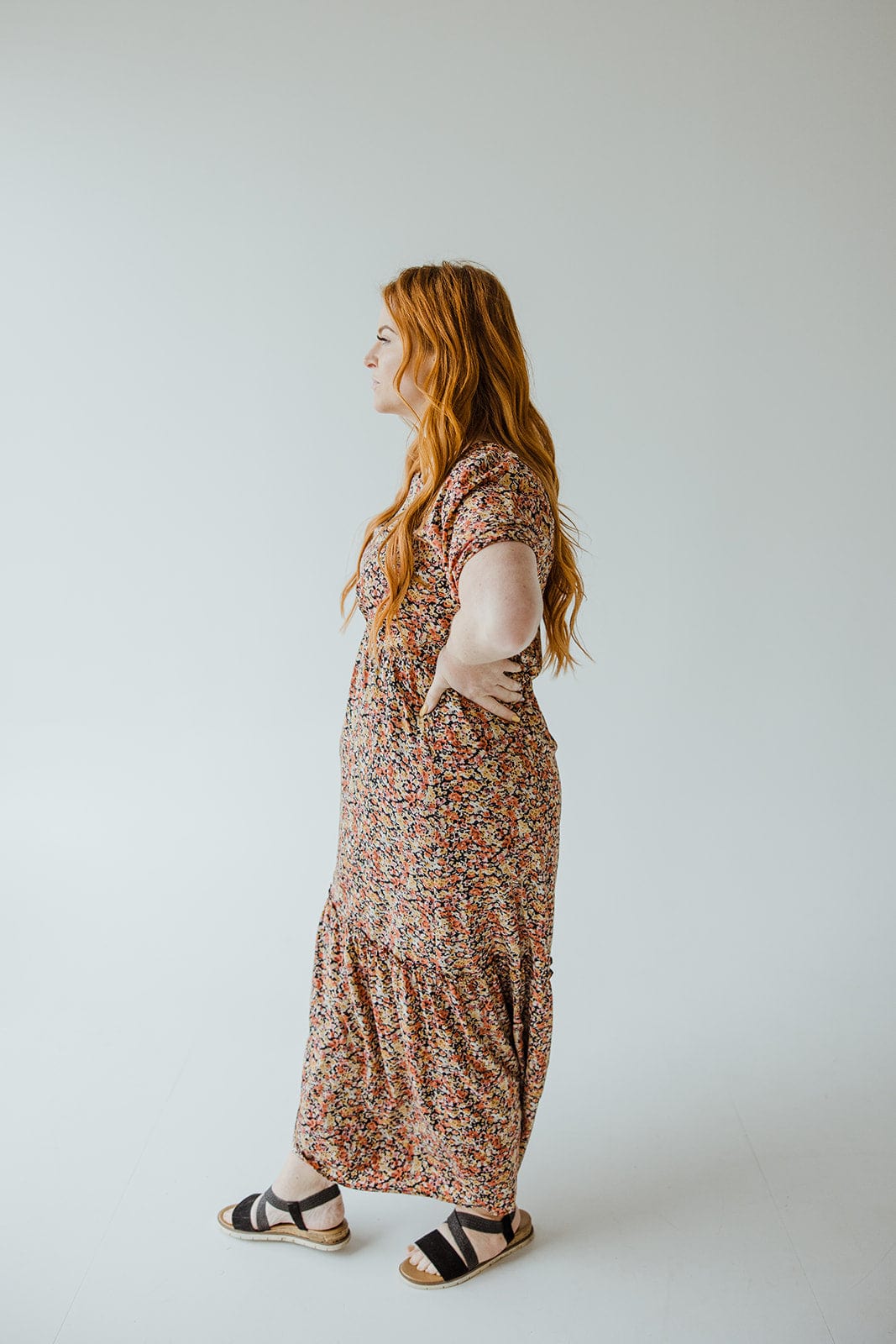 A person with long red hair is wearing the Mikarose Scoop Neck Midi Length Dress with Kick Hem in Autumn Bouquet, standing in profile against a plain white background.