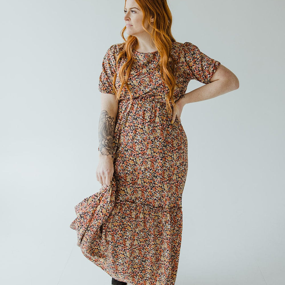 A person with long red hair is wearing a Mikarose scoop neck midi length dress with kick hem in autumn bouquet and black sandals, standing in a neutral pose against a plain background.