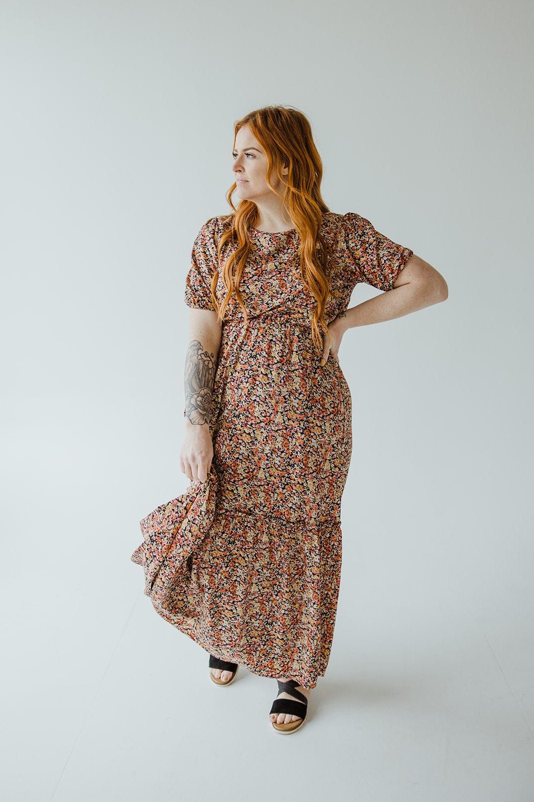 A person with long red hair is wearing a Mikarose scoop neck midi length dress with kick hem in autumn bouquet and black sandals, standing in a neutral pose against a plain background.