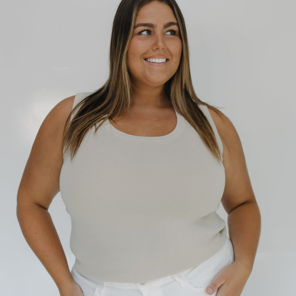 Woman wearing a beige tank top and white pants against a plain background