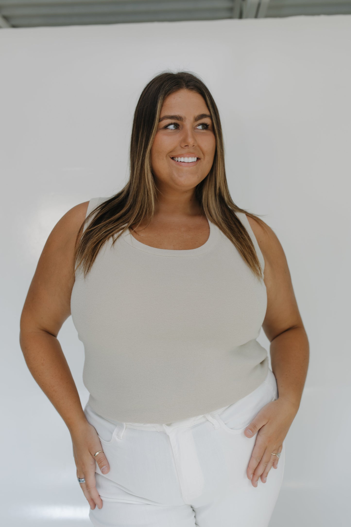 Woman wearing a beige tank top and white pants against a plain background