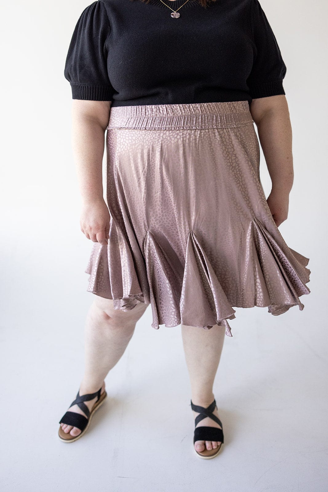 A person wearing a black top, a dusty rose tiered mini skirt, and black sandals stands against a plain background.