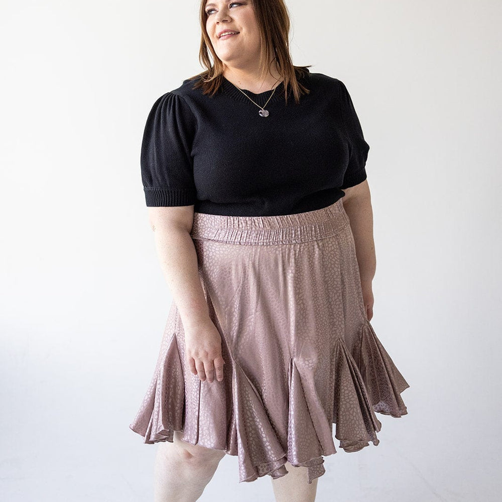 A person in a black top and tiered mini skirt stands on a white background, the dusty rose fabric catching the light as they look to the side.