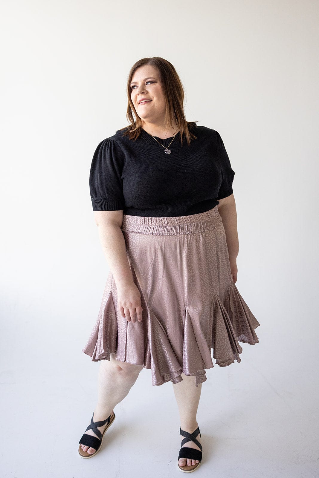 A person in a black top and tiered mini skirt stands on a white background, the dusty rose fabric catching the light as they look to the side.