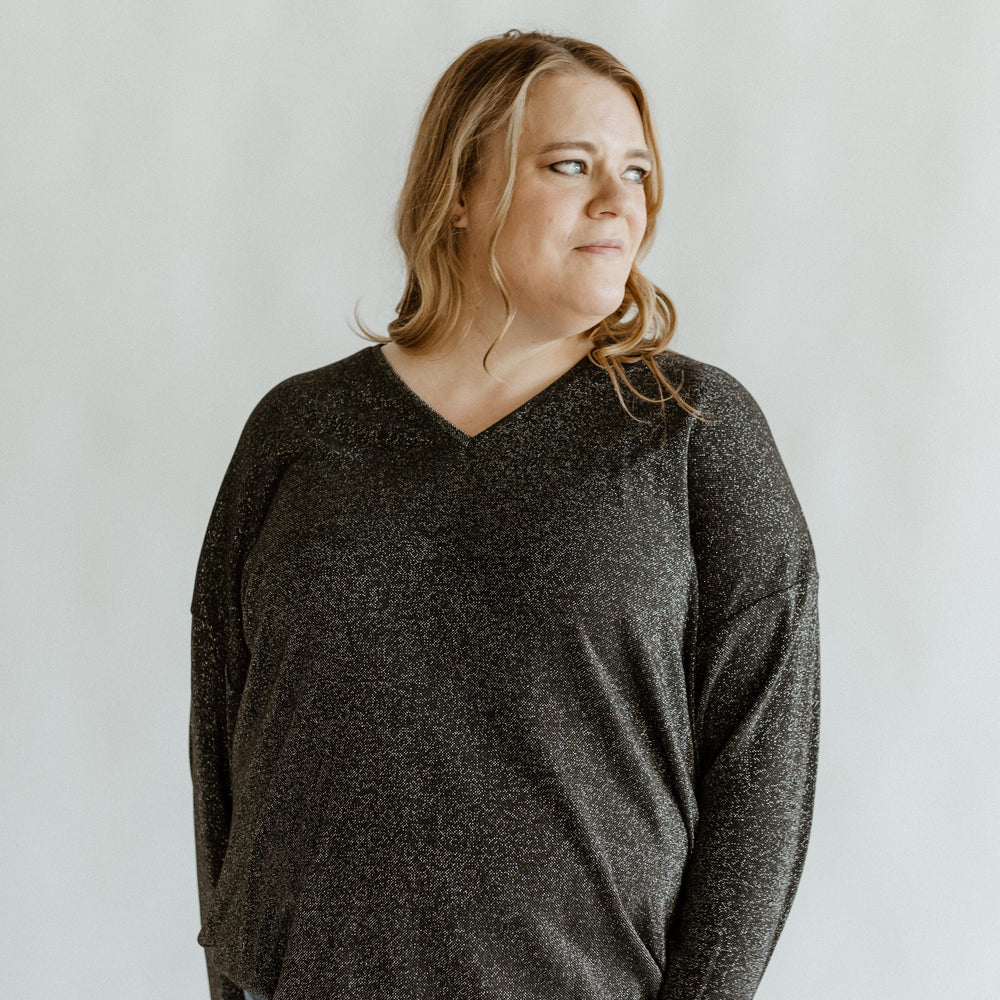 A person with long hair wearing a shimmery banded hem shirt and jeans looks to the side against a plain background.