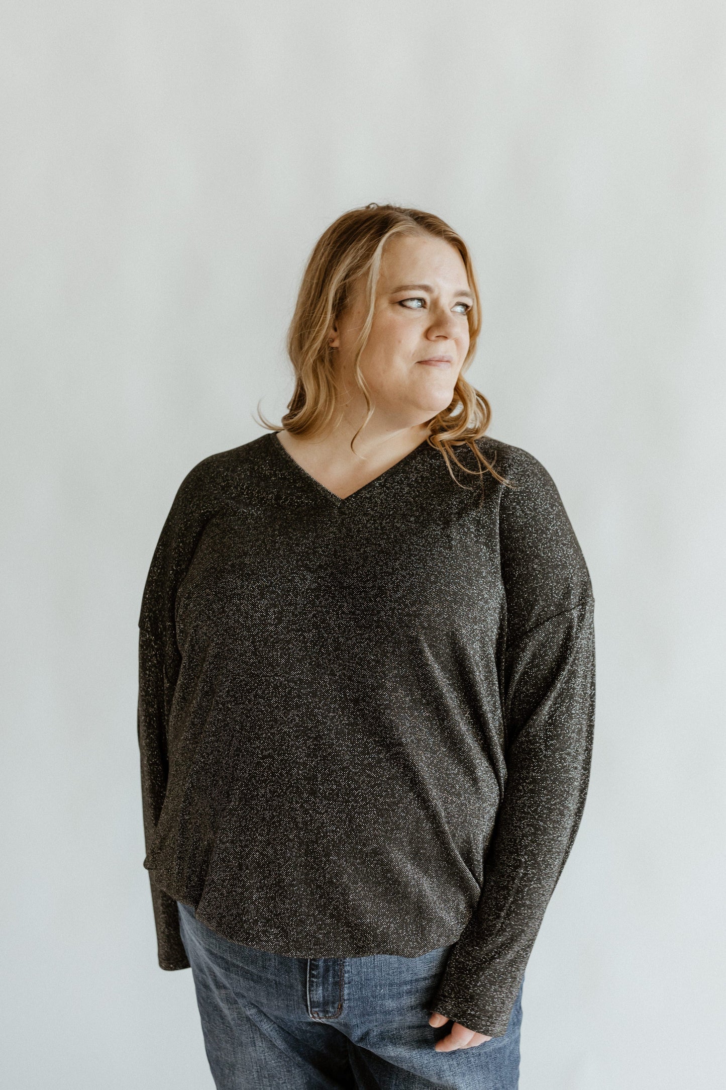 A person with long hair wearing a shimmery banded hem shirt and jeans looks to the side against a plain background.