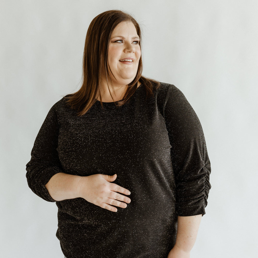 A woman wearing a black 3/4 ruched sleeve blouse with a round neckline, standing against a neutral background.