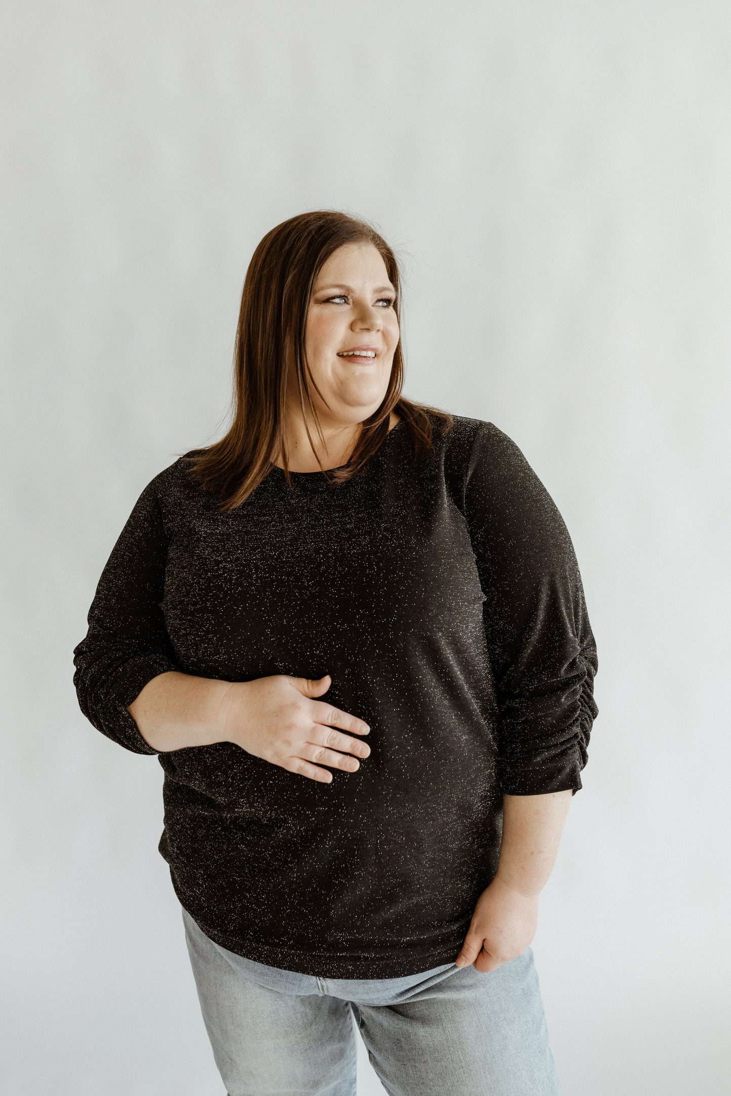 A woman wearing a black 3/4 ruched sleeve blouse with a round neckline, standing against a neutral background.