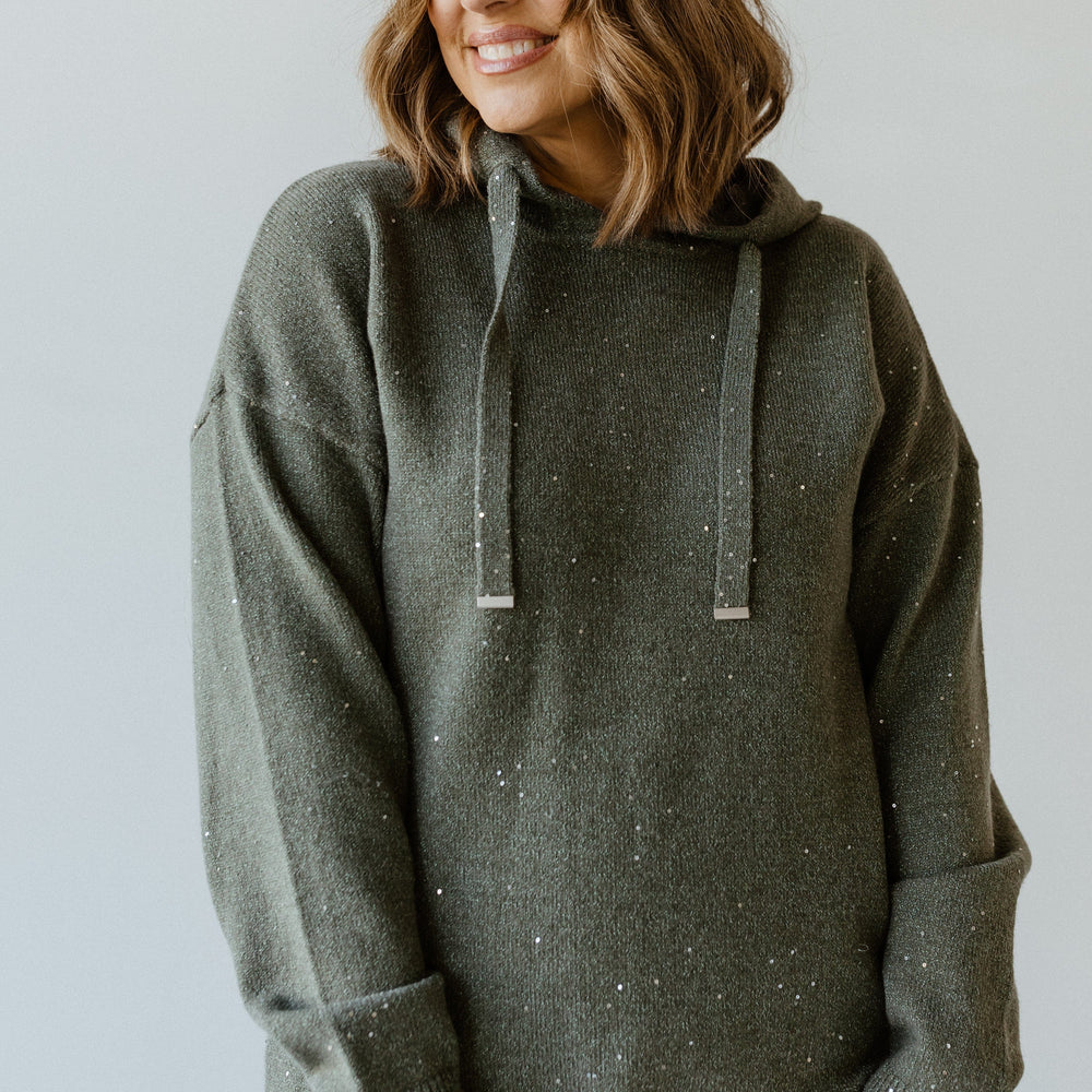 A person with shoulder-length hair is wearing a dark green speckled hoodie with silver toggles and black pants, standing against a light background, smiling and looking to the side.